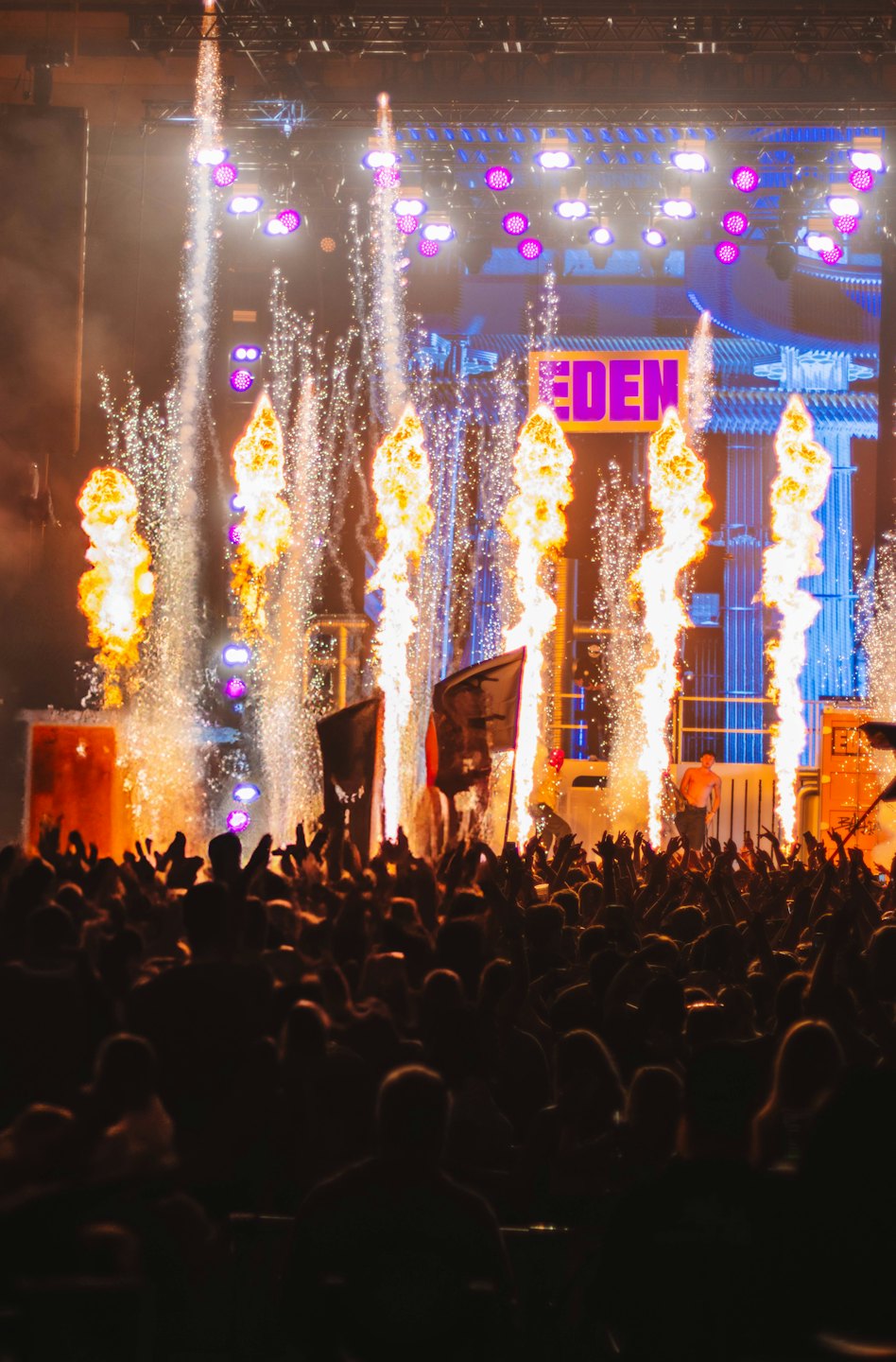 Concert crowd cheering as pyrotechnics erupt from a stage.