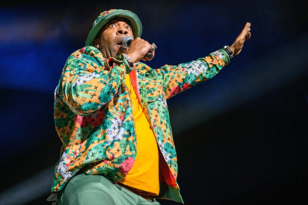 Busta Rhymes performs in a colorful jacket and bucket hat, holding a microphone and raising one hand.