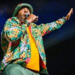 Busta Rhymes performs in a colorful jacket and bucket hat, holding a microphone and raising one hand.