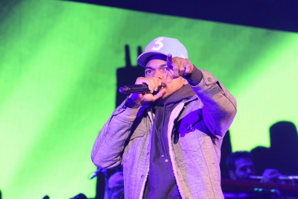 Chance the Rapper performs onstage in a gray jacket and white cap, holding a microphone and gesturing.