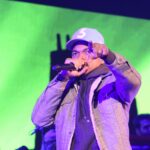 Chance the Rapper performs onstage in a gray jacket and white cap, holding a microphone and gesturing.